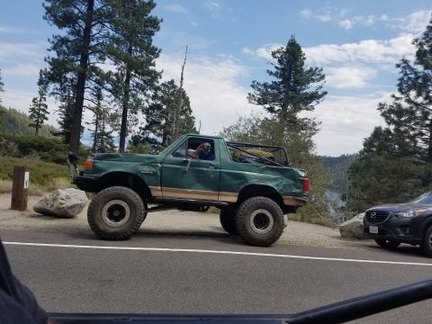 1968 Ford Bronco - A lifted, dark green Ford Bronco with large off-road tires is parked on a dirt pull-off next to a paved road. The vehicle has a tan accent…