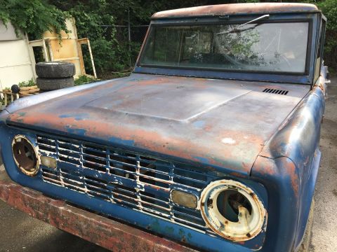 1967 Ford Bronco - The front end of a weathered, blue Ford Bronco is shown in an outdoor setting. The vehicle exhibits significant rust and missing parts, inc…