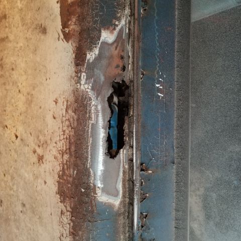 1967 Ford Bronco - A close-up shot shows significant rust and a hole in the blue painted metal of what appears to be a vehicle's body panel. The damaged area…