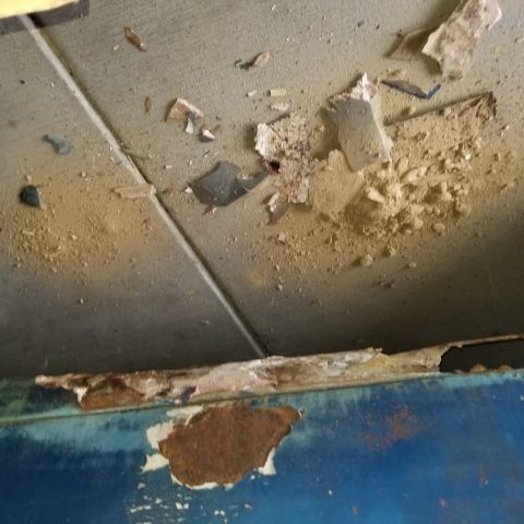 1967 Ford Bronco - Close-up view of heavily rusted and peeling metal trim above a blue painted surface, with debris scattered on a textured ceiling.