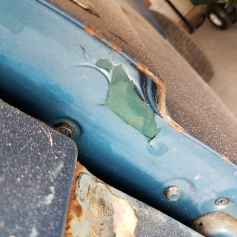 1967 Ford Bronco - Close-up view of peeling blue paint and rust on the edge of a vehicle's body panel. A bolt is visible securing two metal pieces together.