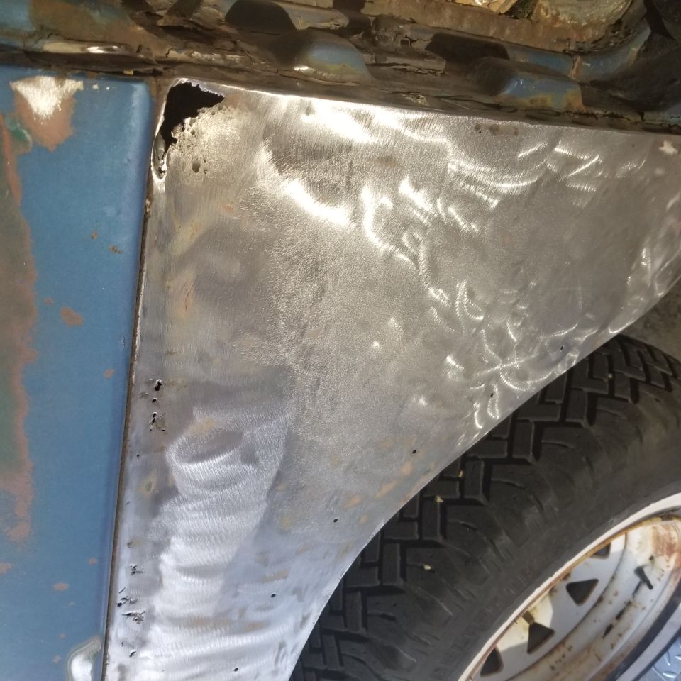 Close-up of a metal patch on a blue vehicle's fender with visible sanding marks and rust.
