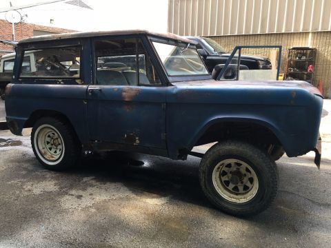 1967 Ford Bronco - The side profile of a weathered, dark blue 1967 Ford Bronco is visible, showing significant rust and damage. The vehicle is parked outdoors…