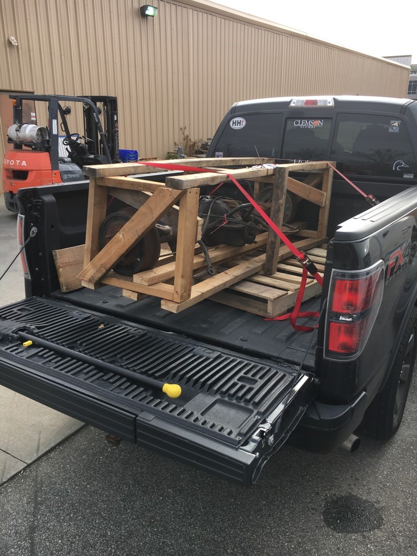 Black Ford F-150 tailgate open with wooden crate of parts secured by red straps. Forklift in background.