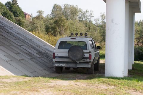 1993 Ford Bronco - The rear of a silver Ford Bronco is parked next to large white pillars, with a wooded area and a building in the background. The vehicle ha…