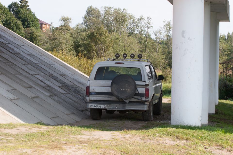 Rear view of a silver Ford Bronco with a spare tire and roof rack lights, parked under pillars.