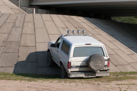 1993 Ford Bronco - A white, two-door Ford Bronco is parked on a concrete ramp under an overpass. The vehicle has a spare tire mounted on the rear and four aux…