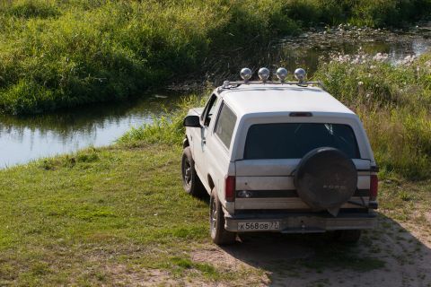 1993 Ford Bronco - The rear view of a white Ford Bronco is parked on a grassy bank next to a body of water. The vehicle has a spare tire mounted on the back a…