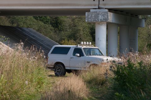 1993 Ford Bronco - A white Ford Bronco is parked under a concrete overpass, surrounded by tall grasses and brush. The vehicle has four auxiliary lights mounte…