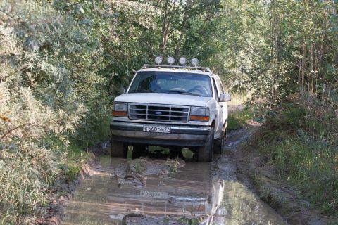 1993 Ford Bronco - A white Ford Bronco is driving through a muddy trail surrounded by trees. The vehicle has four auxiliary lights mounted on the roof rack.