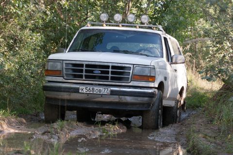 1993 Ford Bronco - A white Ford Bronco is driving through a muddy trail surrounded by trees. The vehicle has four auxiliary lights mounted on a roof rack.
