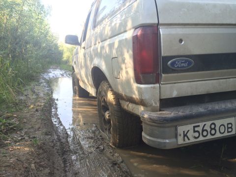 1993 Ford Bronco - The rear-left side of a light-colored Ford Bronco is shown driving through a muddy, water-filled dirt road surrounded by trees. The license…