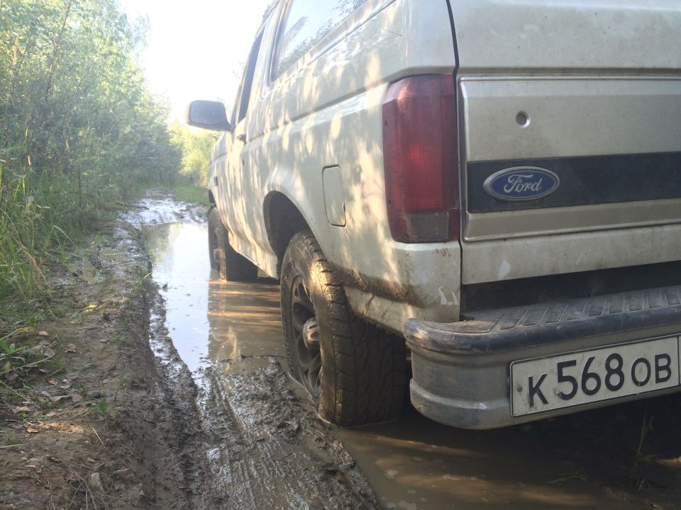 Rear-left view of a light-colored Ford Bronco driving through mud and water on a dirt road.