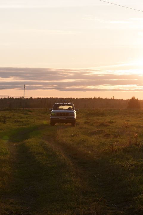 1993 Ford Bronco - A dark-colored SUV is parked in a grassy field during sunset. The vehicle is facing the camera, with the sun setting behind it and casting…