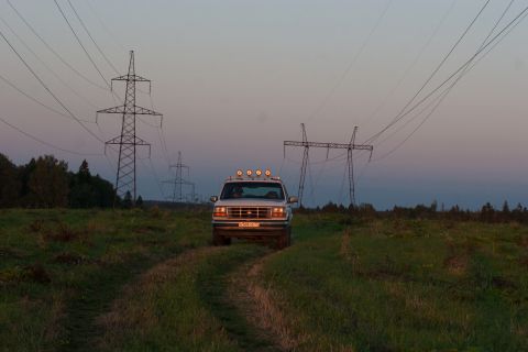 1993 Ford Bronco - A silver Ford Bronco drives on a dirt path through a grassy field at dusk, with large power line towers in the background.