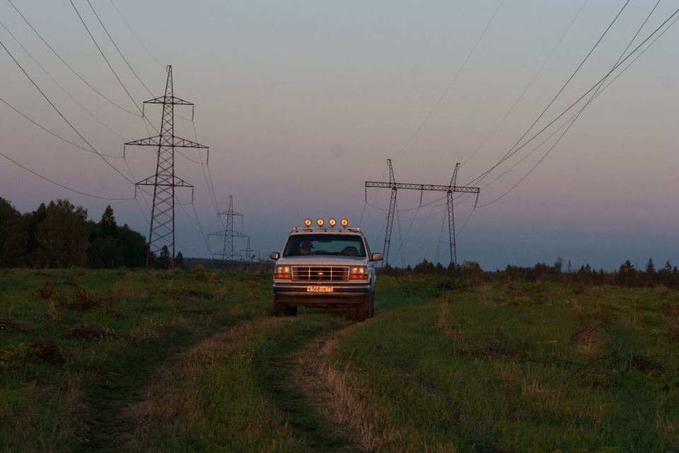 Silver Ford Bronco driving on a dirt path in a field with power line towers at dusk.