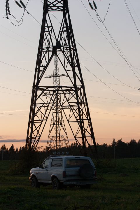 1993 Ford Bronco - A silver Ford Bronco is parked in a grassy field at dusk, with large power line towers silhouetted against the orange sky.