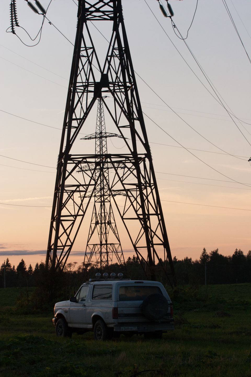Silver Ford Bronco parked in a field with power line towers at sunset.