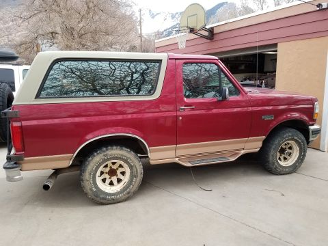 1995 Ford Bronco - The side view of a red and tan 1995 Ford Bronco parked on a concrete driveway next to a garage. The vehicle has off-road tires and a spare…