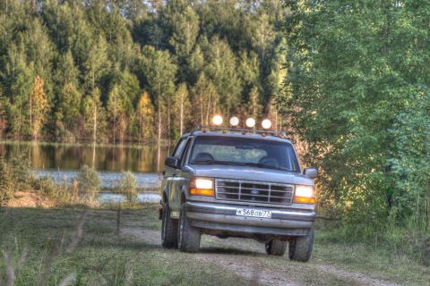 1993 Ford Bronco - A silver 1993 Ford Bronco is parked on a dirt road next to a lake with a forest in the background. The SUV has four auxiliary lights mounte…