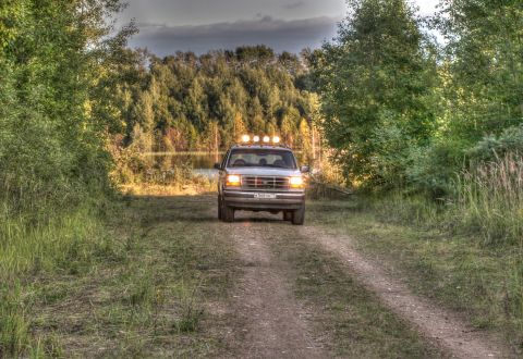 1993 Ford Bronco - A white Ford SUV with four auxiliary lights mounted on the roof rack drives down a dirt path towards a lake surrounded by trees. The vehicl…