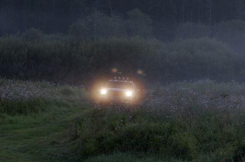 1993 Ford Bronco - A white Ford Bronco with its headlights and roof-mounted lights on drives through a grassy field at dusk. Tall trees and foliage form a dar…