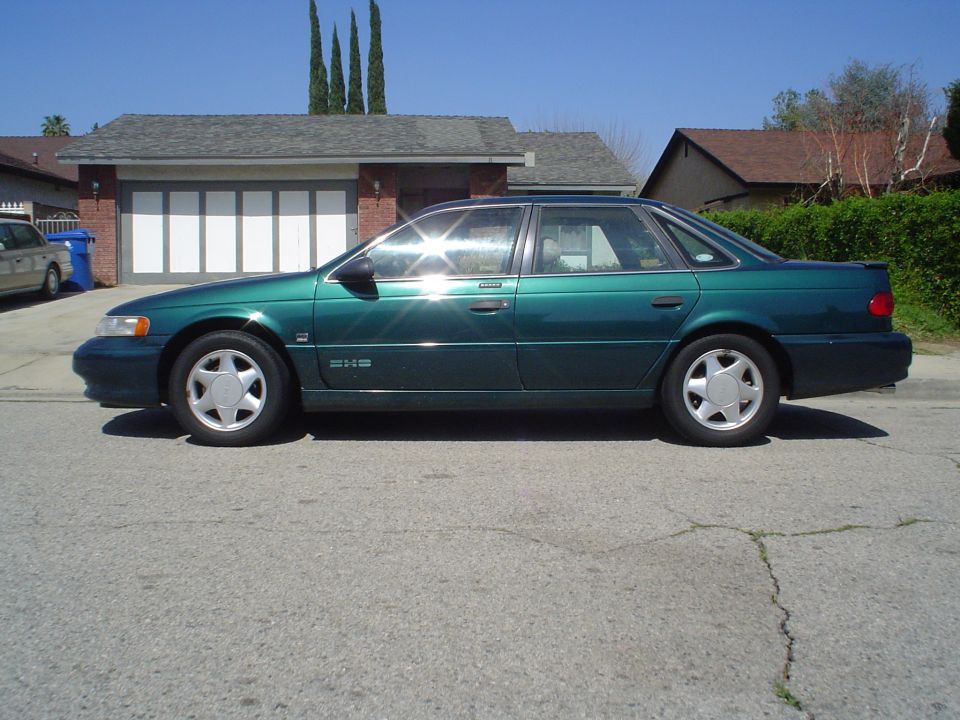Green Ford Taurus SHO in profile parked on a street before a brick house and garage