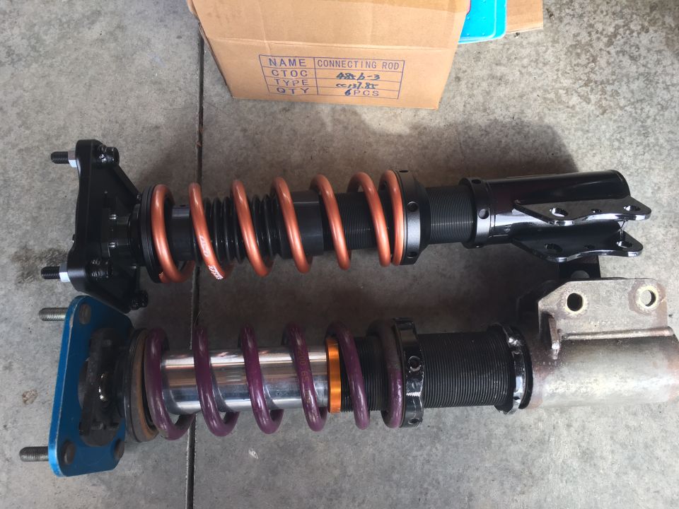 Two coilover suspension assemblies, one with copper springs and one with purple springs, sit on a concrete surface next to a cardboard box