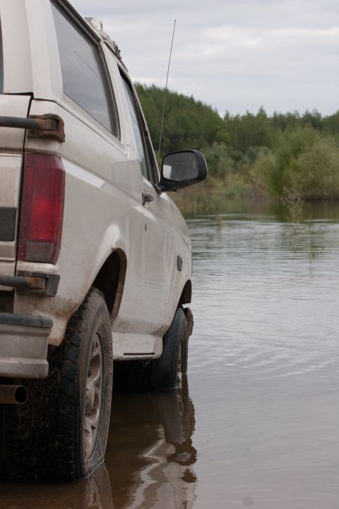 1993 Ford Bronco - A white 1993 Ford Bronco is seen from the side, partially submerged in shallow water with trees in the background.