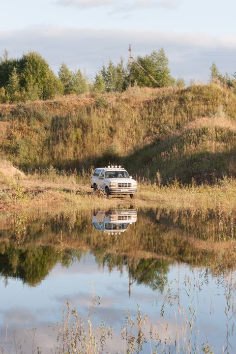 1993 Ford Bronco - A white SUV sits near the edge of a calm lake, its reflection perfectly mirrored in the water.