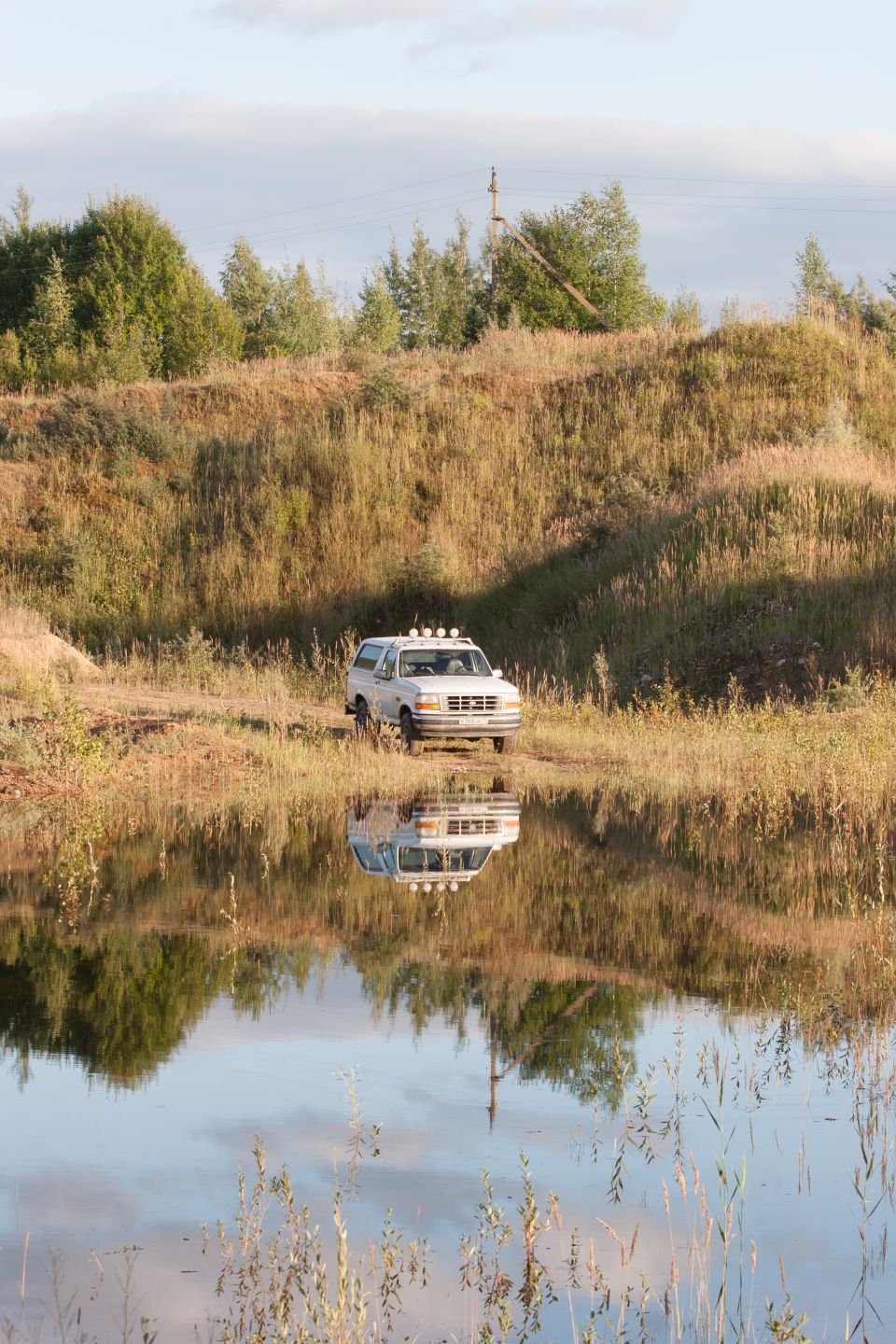 White SUV with roof lights parked by a body of water, reflecting in the water