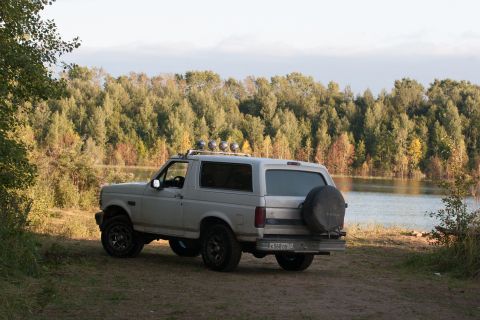 1993 Ford Bronco - A silver Ford Bronco is parked on a dirt path next to a lake with trees in the background.