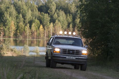 1993 Ford Bronco - This 1993 Ford Bronco, equipped with a roof-mounted light bar, is captured driving on a dirt path adjacent to a tranquil lake.