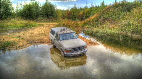 1993 Ford Bronco - A light-colored Ford Bronco navigates a water crossing surrounded by natural terrain.