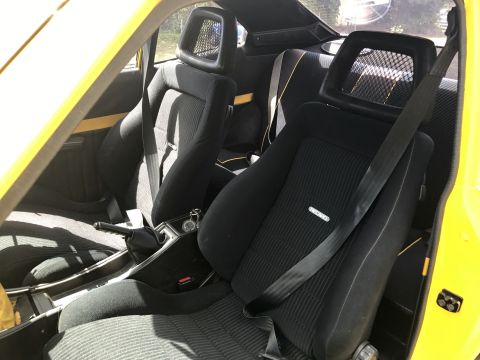 1973 Mazda RX-3 - The interior of a yellow car features black fabric seats with distinctive headrests and seatbelts. A Recaro logo is visible on the passenge…