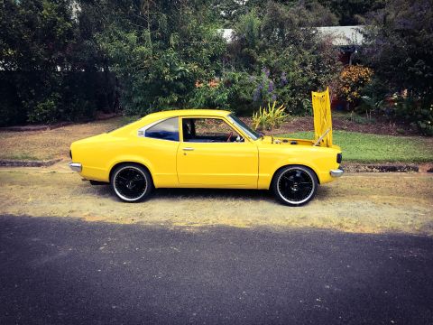 1973 Mazda RX-3 - A yellow 1973 Mazda RX-3 coupe is parked on the side of a street with its hood open, showing custom black rims.