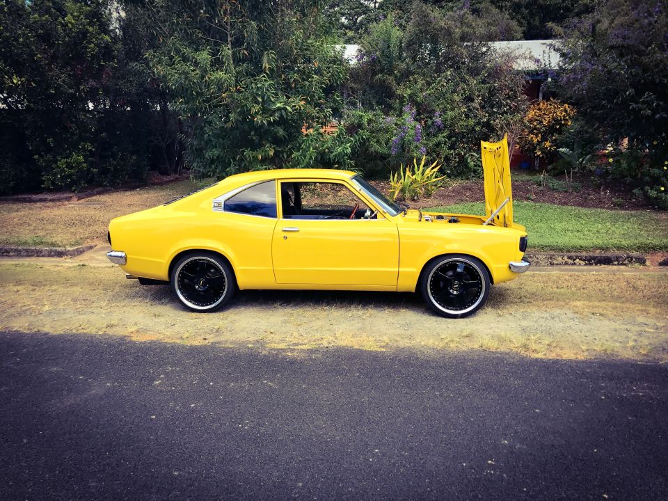 Yellow 1973 Mazda RX-3 coupe with hood open, side view, black rims, parked on a street