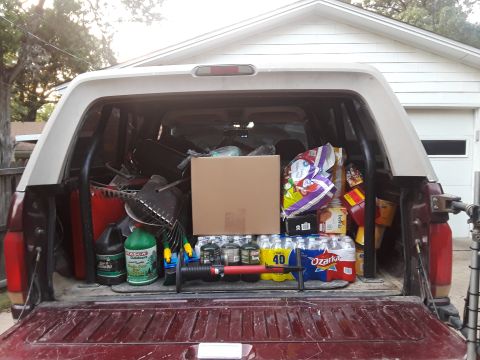 1996 Ford Bronco - The bed of a pickup truck is packed with various items, including bottled water, snacks, gardening tools, and a large cardboard box.