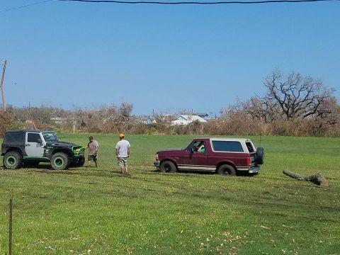 1996 Ford Bronco - Two off-road vehicles are parked in a field under a clear blue sky, with a few houses and trees in the background.