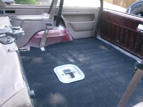1996 Ford Bronco - The interior cargo area of a vehicle features dark carpeting and a square metal access panel on the floor.