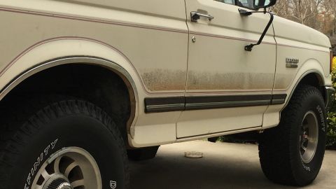1988 Ford Bronco - The side of a white Ford Bronco shows dirt streaks and a red pinstripe above the black trim.