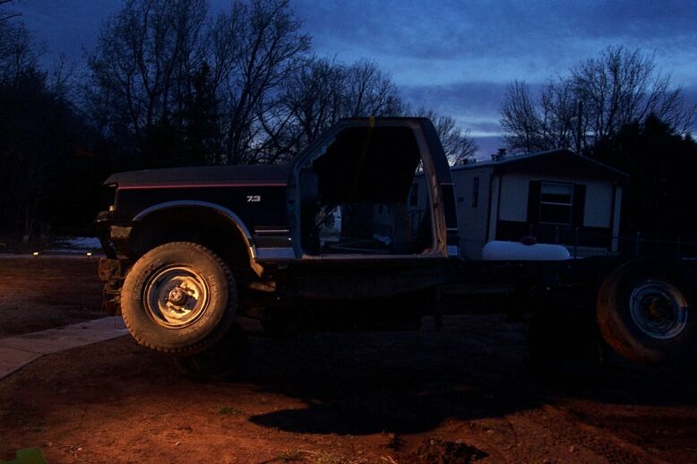 Stripped 1980s Ford 4x4 project on its bare frame at dusk, cab doorless with a 7.3 fender badge