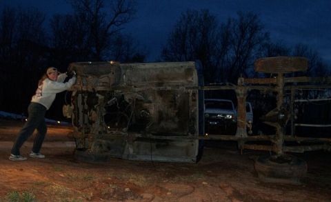 1990 Ford Bronco - A person attempts to move a heavily damaged, overturned vehicle chassis at dusk. Another vehicle is in the background.
