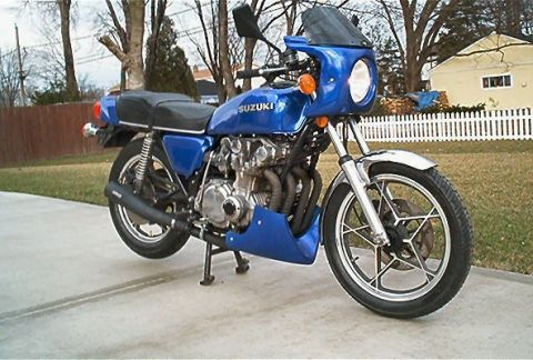 1985 Ford Crown Victoria LTD - A blue Suzuki motorcycle with a small windscreen is parked on a concrete driveway in front of a white picket fence.
