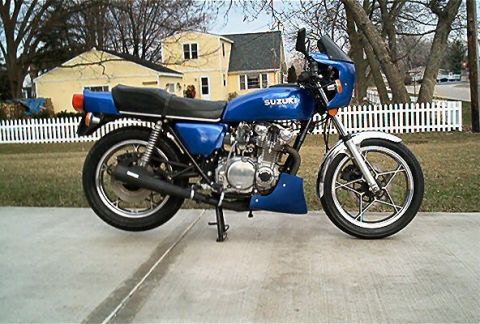 1985 Ford Crown Victoria LTD - A blue Suzuki motorcycle is parked on a concrete driveway in front of a white picket fence and a yellow house.