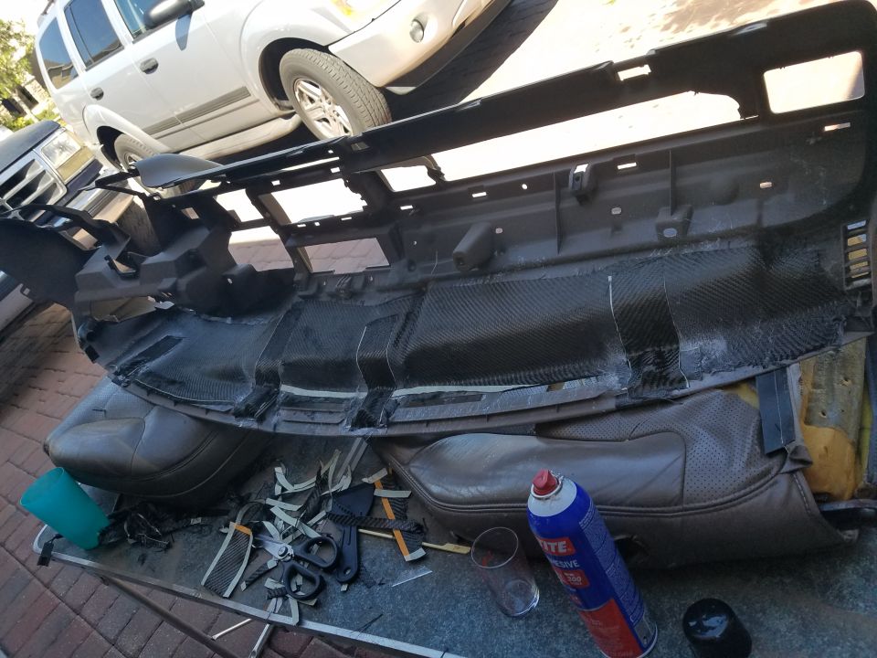 Car dashboard disassembled on a table, with parts and tools scattered around. A white SUV is parked behind it