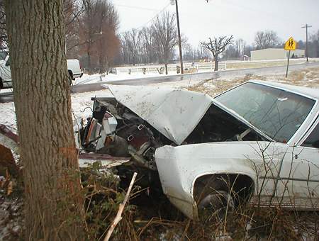 1989 Ford Crown Victoria - A white sedan lies wrecked after colliding with a tree, its front end severely damaged and exposed on a cold, snowy day.