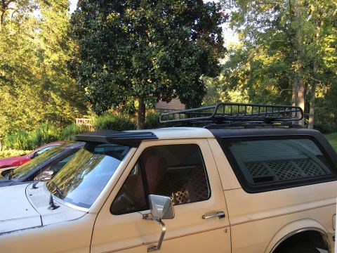 1988 Ford Bronco - A cream-colored Ford Bronco is parked next to a red car, with trees and a house in the background.