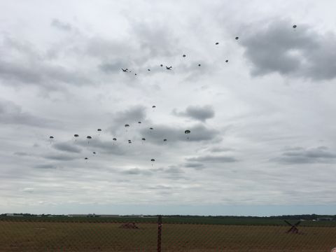 1996 Ford Bronco - A formation of paratroopers descends from aircraft under a cloudy sky, with a field and fence in the foreground.