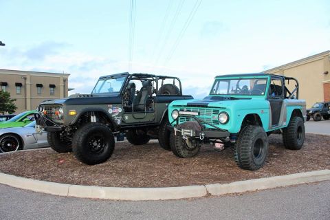1972 Ford Bronco - Two modified Ford Broncos, one olive green and one teal, are parked outdoors with a building and a green sports car visible.